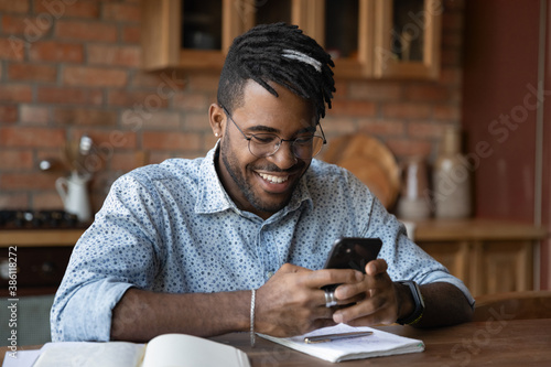 Smiling 20s African American man sit at home kitchen office text message on smartphone online. Happy young biracial male in glasses use cellphone browse surf wireless internet on modern gadget.