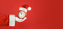 Santa Claus Hand Holding Red Alarm Clock In Santa Hat. Red Background. Concept Of Coming Christmas And New Year, Holiday Sales. Space For Text, Banner