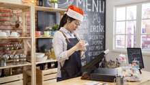 Small Business Owner Holding Notebook At Counter In Coffee Shop. Asian Female Barista In Red Santa Hat Writing Note At Bar In Cafe. Girl Server In Coffeehouse Celebrating Xmas Holidays In Store.