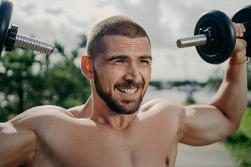 Handsome muscular unshaven man with bristle raises dumbbells, does exercises and lifts barbells, stands with naked body outdoor. Strong bodybuilder makes weightlifting, poses with sport equipment