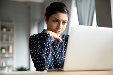 Serious indian businesswoman sitting at desk looking at laptop screen read e-mail thinking, search issue solution feel worried. Student and difficult task, hard test exam, unmotivated employee concept