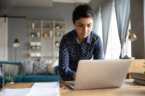 Focused serious indian female entrepreneur standing bent over device typing at laptop finishing task in hurry, working on project solve problems remotely. Modern wireless e technologies usage concept