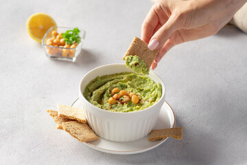 Female hand dipping cracker into white bowl with avocado hummus. healthy vegan dip. close-up. gray background with place for text.