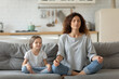 Peaceful young mother and little daughter meditating, doing yoga exercise, sitting in lotus pose with mudra gesture on cozy couch in living room, family enjoying leisure time, relaxing together