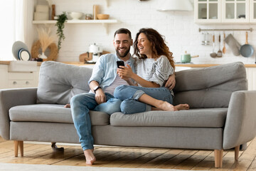 happy young woman and man hugging, using smartphone together, sitting on cozy couch at home, smiling