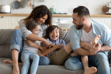 Happy Young Parents With Two Kids Having Fun At Home, Laughing Overjoyed Mother And Father Tickling Little Daughter And Son, Sitting On Couch In Living Room, Family Enjoying Leisure Time Together