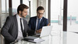 © fizkes - Focused young male business partners in suits sitting at table, looking at computer screen, discussing corporate software, finding problem solution or developing online project ideas at meeting.