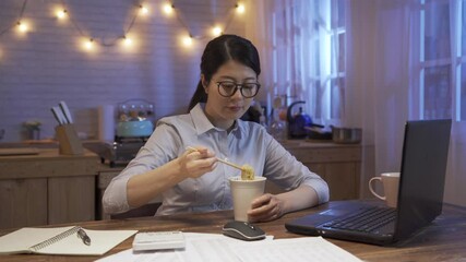 Wall Mural - asian korean office lady in glasses working on laptop computer and using chopsticks to eat tasty ramen soup in disposable cup in kitchen at night