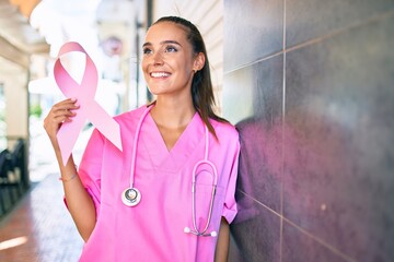 Wall Mural - Young doctor woman holding pink cancer ribbon leaning on the wall at street of city.