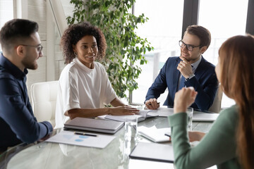 Wall Mural - Happy european leader with multiracial colleagues listening to smiling african american female coworker, sharing profitable successful project ideas at brainstorming meeting, sitting together at table