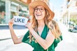 © Krakenimages.com - Young beautiful blonde woman on vacation wearing summer hat smiling happy. Standing with smile on face holding paper with like message at street of city.