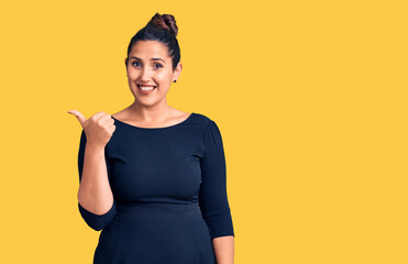 Young beautiful brunette woman wearing casual dress smiling with happy face looking and pointing to the side with thumb up.