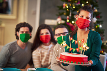 Wall Mural - Group of friends in masks celebrating Christmas at home with fancy cake