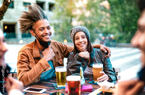 Young couple drinking beer glasses with open face masks - New normal lifestyle concept with friends having fun together on happy hour at brewery bar - Bright vivid filter with focus on girl with hat