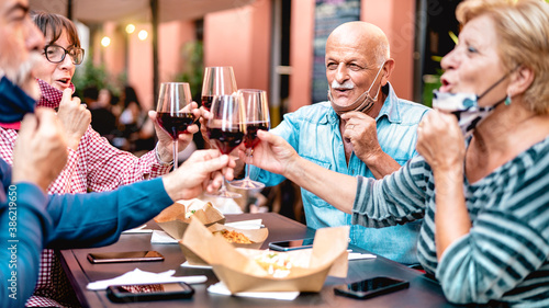 Senior friends toasting wine at restaurant bar wearing opened face mask - New normal lifestyle concept with happy mature people having fun together at garden party - Warm filter with focus on bald man