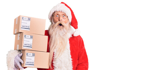 Wall Mural - Old senior man with grey hair and long beard wearing santa claus costume holding boxes scared and amazed with open mouth for surprise, disbelief face