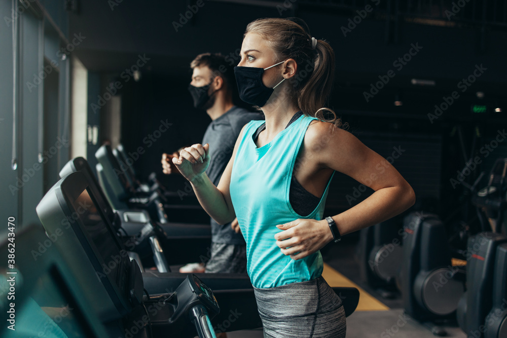 Young fit woman and man running on treadmill in modern fitness gym. They keeping distance and wearing protective face masks. Coronavirus world pandemic and sport theme. - obrazy, fototapety, plakaty 
