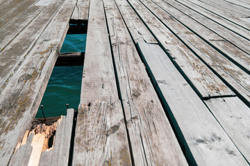 An old wooden dock with a broken board and a hole in the floor where the blue surface of the sea is visible