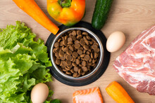 Dry Pet Dog Food With Natural Ingredients. Raw Meat, Fish, Vegetables, Eggs And Salad Near Bowl With Dry Pet Feed On Wooden Background. Concept Of A Correct Balanced And Healthy Nutrition For Pet