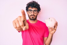 Handsome Young Man With Curly Hair And Bear Holding Piggy Bank Pointing With Finger To The Camera And To You, Confident Gesture Looking Serious