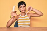 Brunette teenager girl eating banana as healthy snack with angry face, negative sign showing dislike with thumbs down, rejection concept