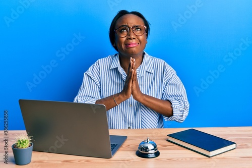 Young african woman working at hotel reception using laptop begging and praying with hands together with hope expression on face very emotional and worried. begging.