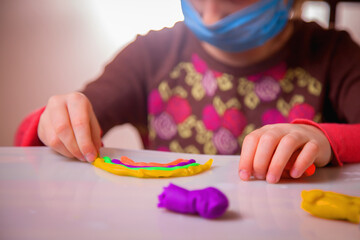 Living under coronavirus quarantine: game and occupation for child at home during quarantine of coronavirus. Young girl with protect medical face mask making plasticine statues of rainbow.