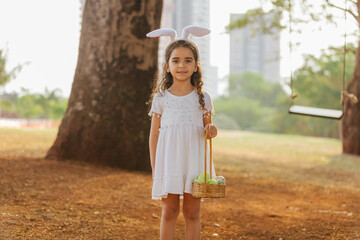 Wall Mural - curly latin girl smiling wearing bunny ears and holding a hamper of easter eggs in the park