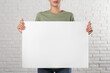 © New Africa - Woman holding blank poster near white brick wall, closeup. Mockup for design