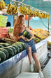 © Nikolay - A young red-haired girl with freckles in a blue dress sits on the counter and holds a watermelon in her hands. Fruit market in Thailand