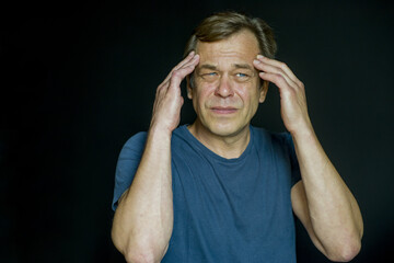 Portrait of a 45-50-year-old man holding his head in his hands on a dark background. He may have frequent headaches or migraines.