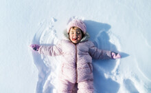 Top View Portrait Of Cheerful Small Girl Lying In Snow In Winter Nature, Making Angels.