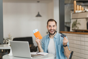 Wall Mural - Excited young man sit alone at table in coffee shop cafe restaurant indoors working or studying on laptop pc computer hold passport tickets credit bank card. Freelance mobile office business concept.