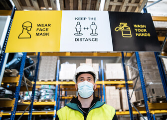 Young worker with face mask standing indoors in warehouse, coronavirus concept.