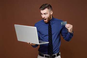 Shocked bearded young business man wearing blue shirt tie working on laptop pc computer hold credit bank card isolated on brown background studio portrait. Achievement career wealth business concept.