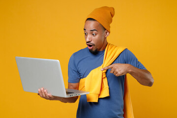 Shocked amazed young african american man 20s wearing casual blue t-shirt hat standing working pointing index finger on laptop pc computer isolated on bright yellow colour background, studio portrait.