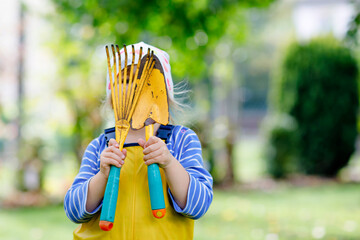 Wall Mural - Adorable little toddler girl holding garden tools in hands. Cute child learn gardening, planting and cultivating vegetables in domestic garden. Kid with shovel and rake. Ecology, organic food.