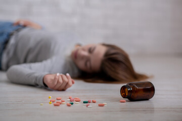 Wall Mural - Young woman lying on floor with bottle of tablets scattered on floor, committing suicide, selective focus