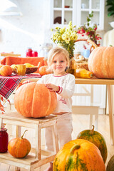 Wall Mural - A little blonde girl sits on a wooden floor in a white kitchen next to a large orange autumn pumpkin