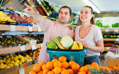 Positive cheerful smiling family couple standing with full cart after shopping and pointing to shelves in fruit store