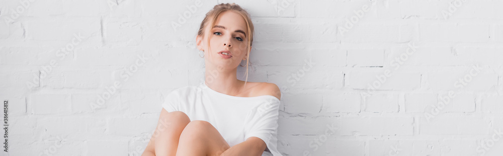 panoramic orientation of young blonde woman looking at camera while sitting near white wall - obrazy, fototapety, plakaty 