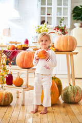 Wall Mural - A little blonde girl sits on a wooden floor in a white kitchen next to a large orange autumn pumpkin