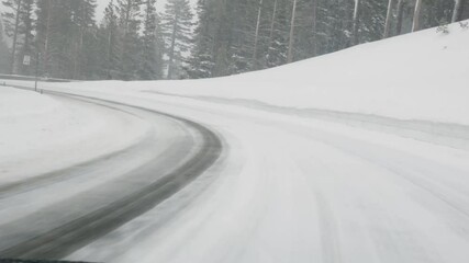 Wall Mural - Driving on road with falling snow. Traffic on snowy icy mountain road with pine trees. Danger travel conditions in winter, 4k