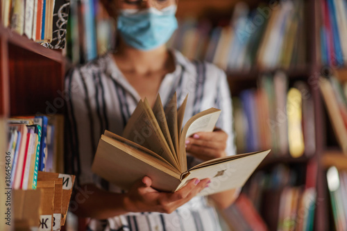 Closeup of attractive college girl standing in library with face mask on and reading a book. Studying during corona virus concept.