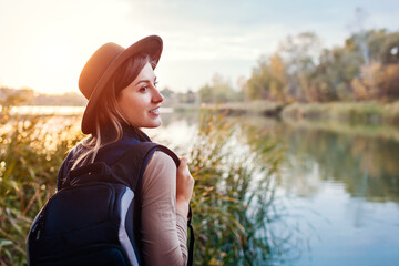 Wall Mural - Traveler with backpack relaxing by autumn river at sunset. Young woman enjoys fall weather and landscape