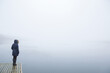 Young adult woman standing alone on edge of footbridge and staring at lake. Mist over water. Foggy air. Early chilly morning. Peaceful atmosphere. Empty place for text, quote or sayings. Back view.