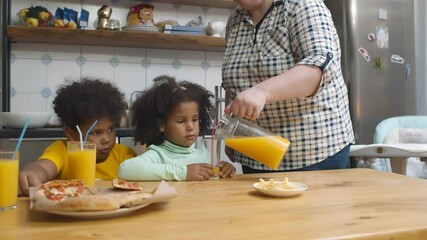 Wall Mural - Caucasian mother pouring juice in glass for mixed race children sitting in kitchen and eating pizza