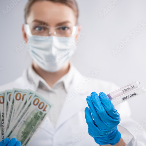 Doctor in white medical gown, protective blue gloves, mask and glasses keeps money and covid-19 vaccine on white background. Scientific discovery reward. Healthcare and coronavirus infection concept.