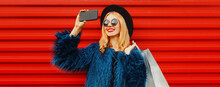 Happy Smiling Blonde Woman Taking Selfie Picture By Smartphone With Shopping Bags Wearing Blue Faux Fur Coat, Black Round Hat Over Red Wall Background