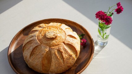 Homemade bread (French bun) on a dark wood plate, in the sun. And a red flower in a glass nearby